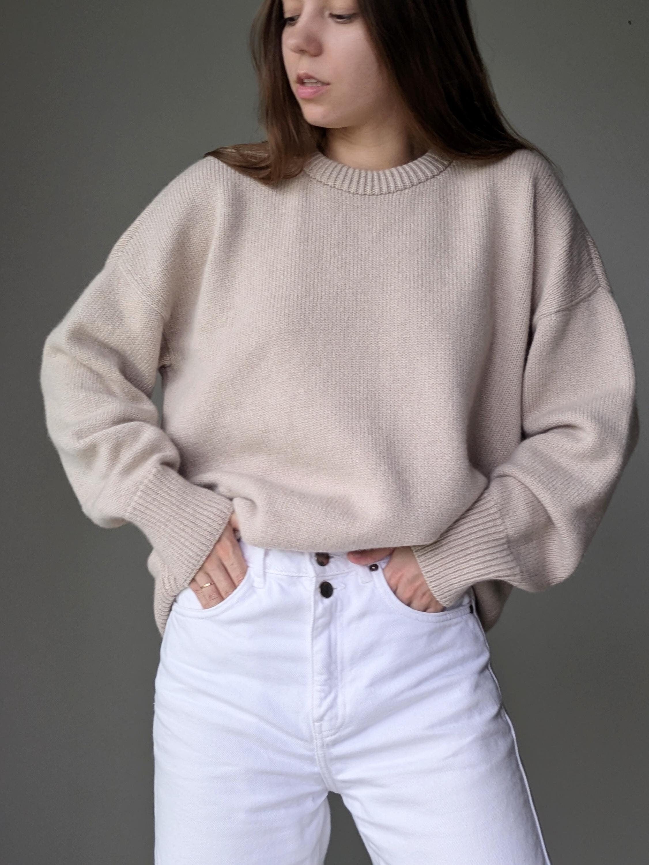 Person wearing a beige sweater and white pants against a gray background