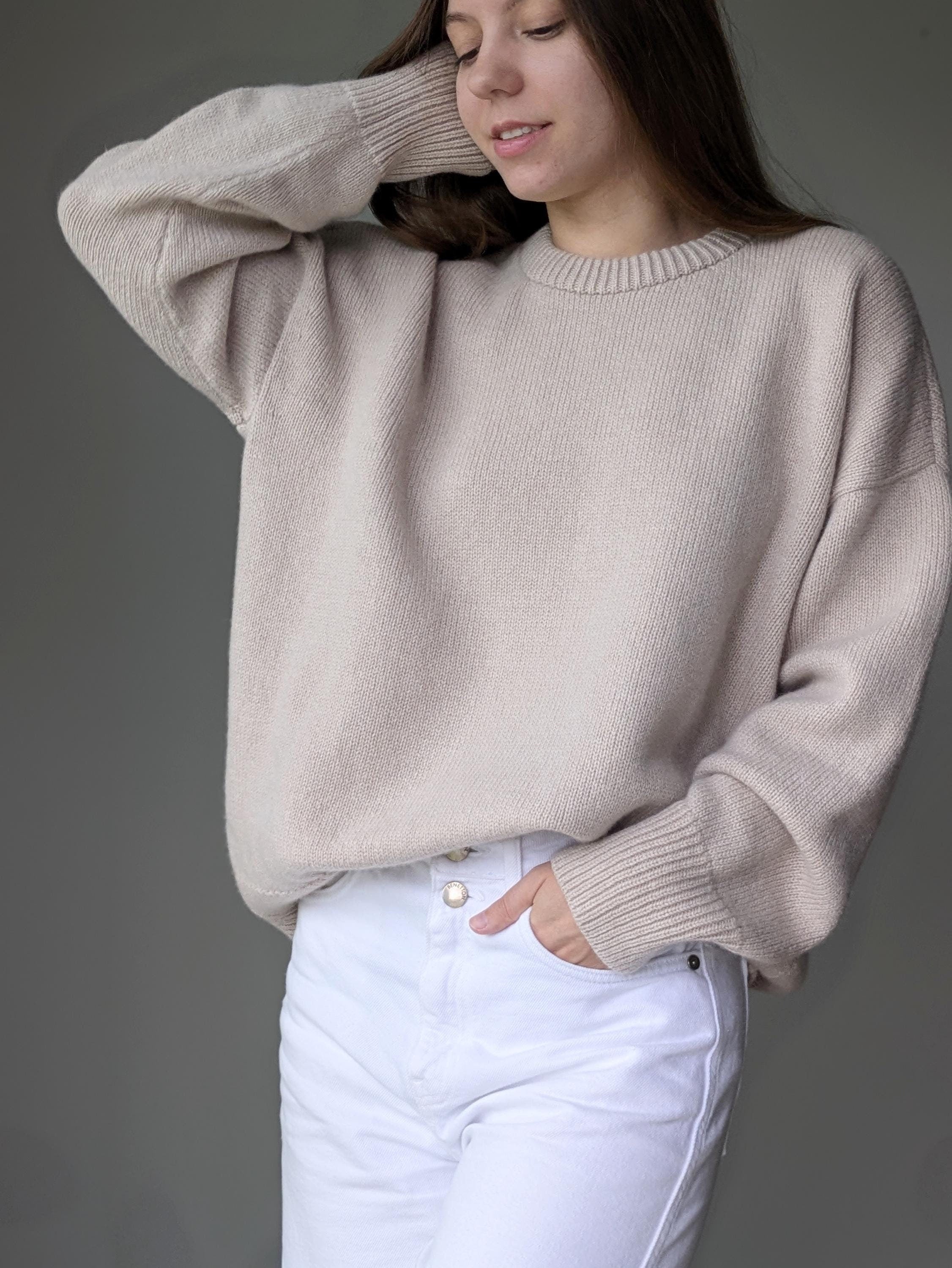 Person wearing a beige sweater and white pants against a gray background