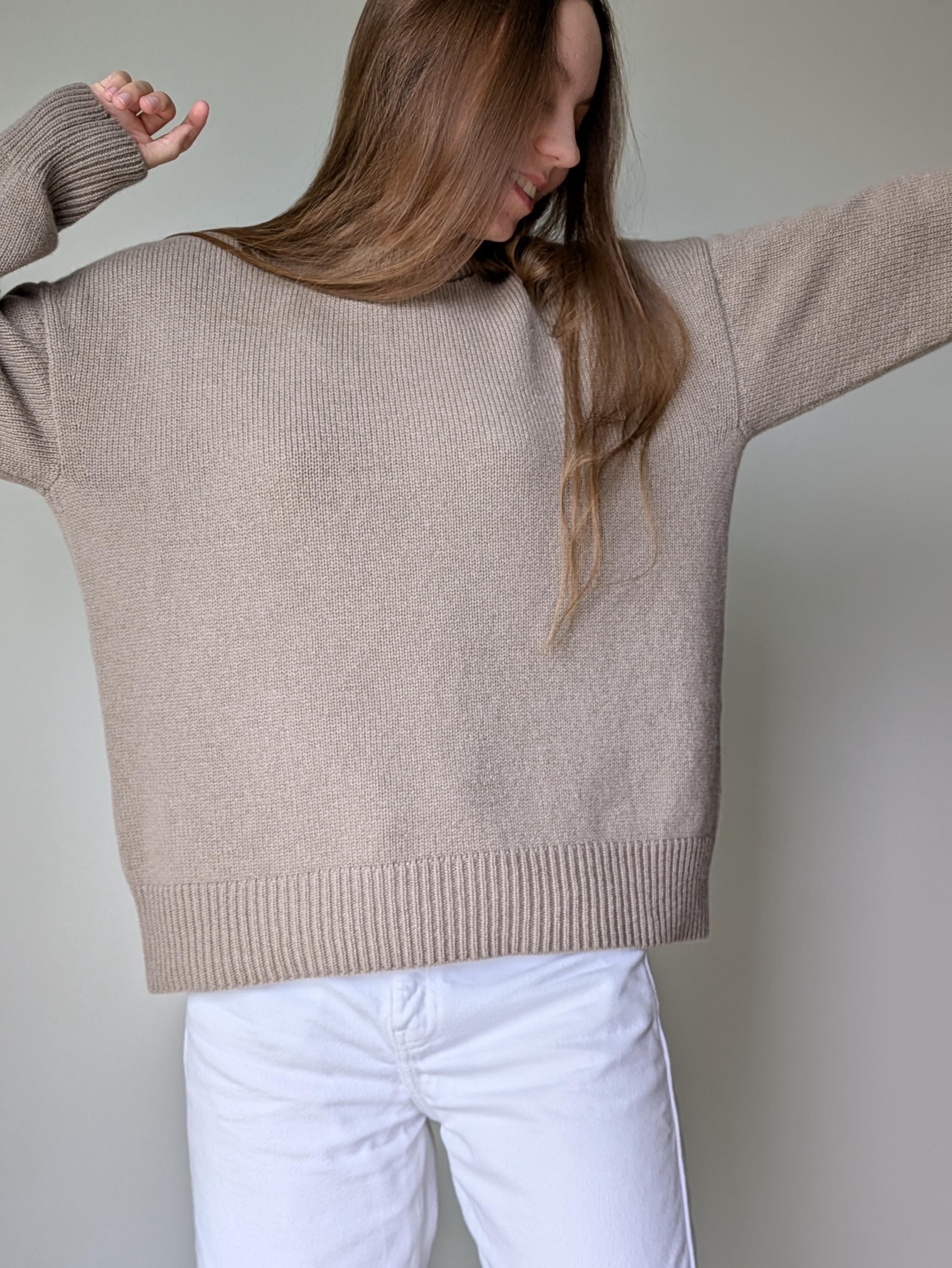Person wearing a beige sweater and white pants against a plain background