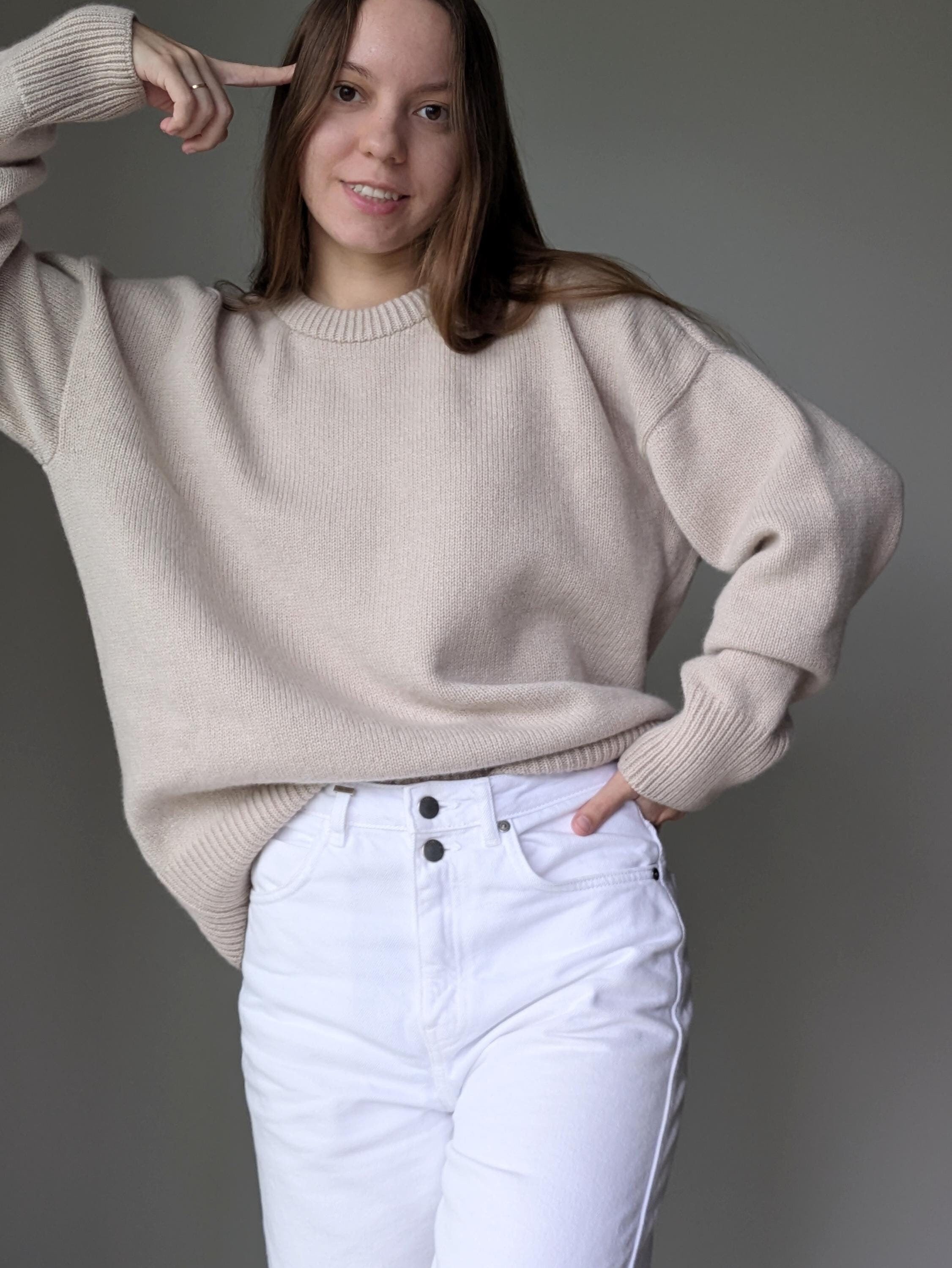 Person wearing a beige sweater and white pants against a gray background