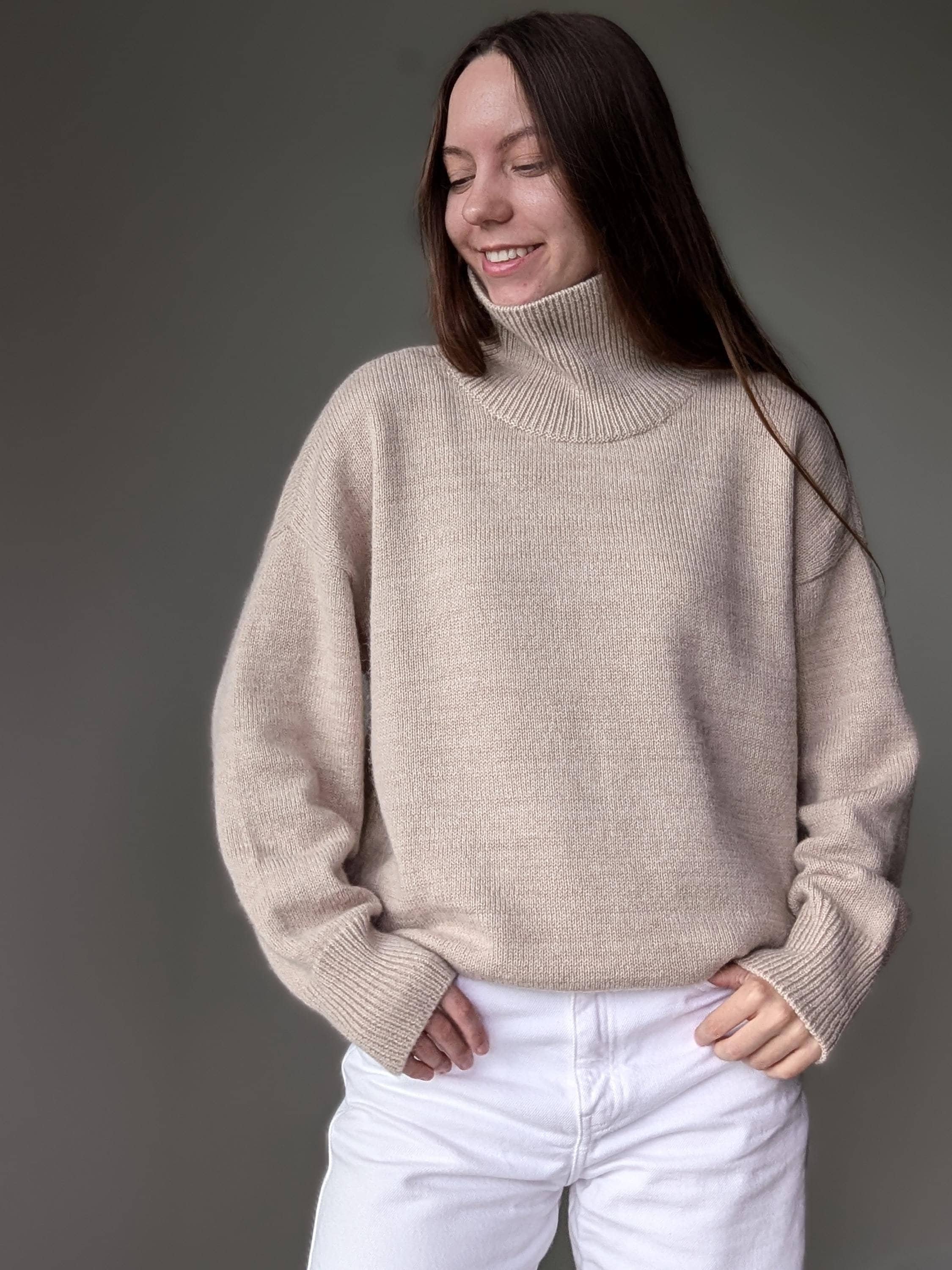 Sand woman wearing a beige sweater and white pants against a gray background