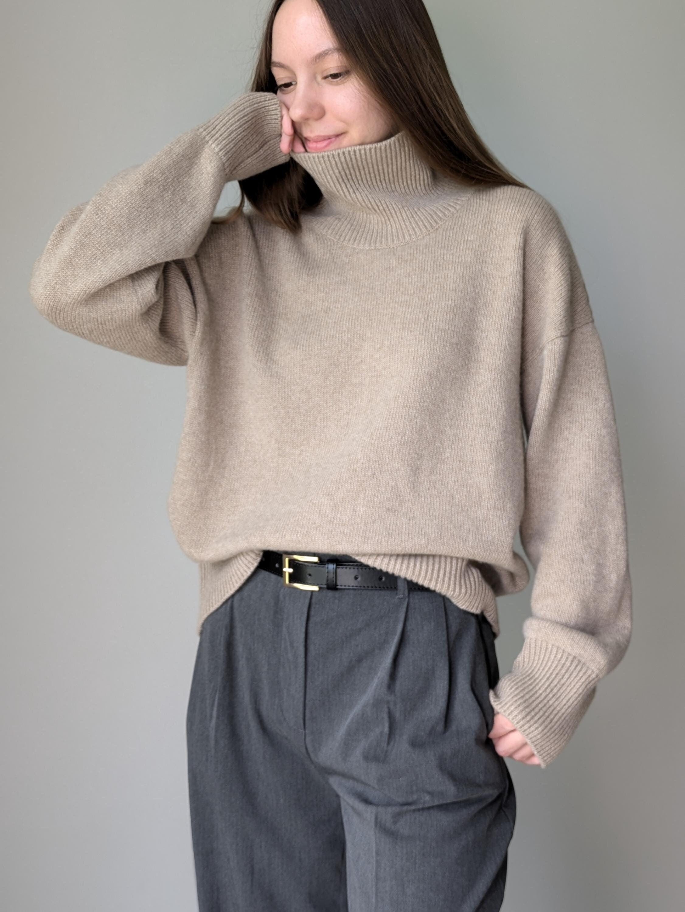 Beige Woman wearing a beige sweater and gray pants against a plain background