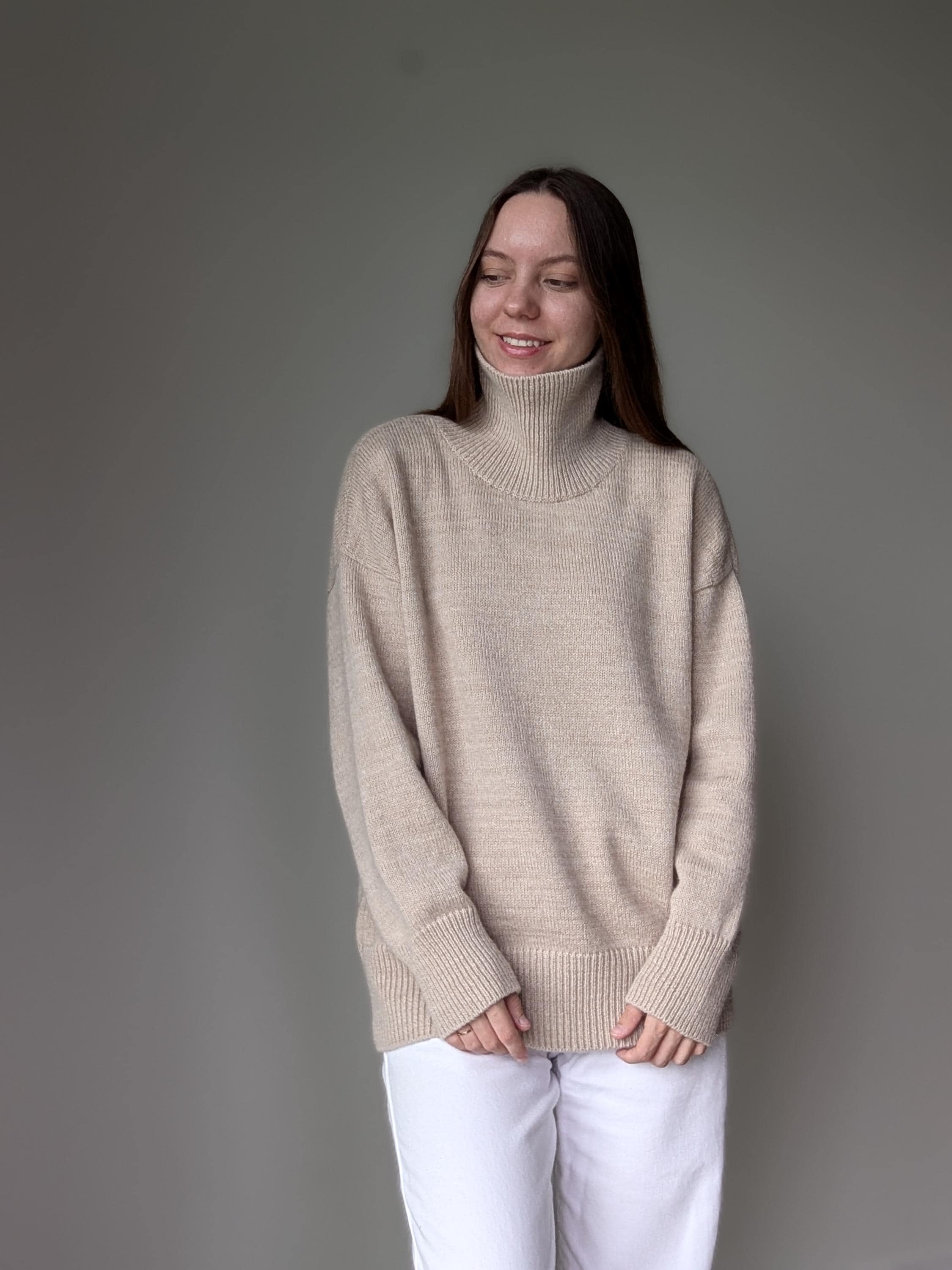 Sand woman wearing a beige turtleneck sweater and white pants against a gray background