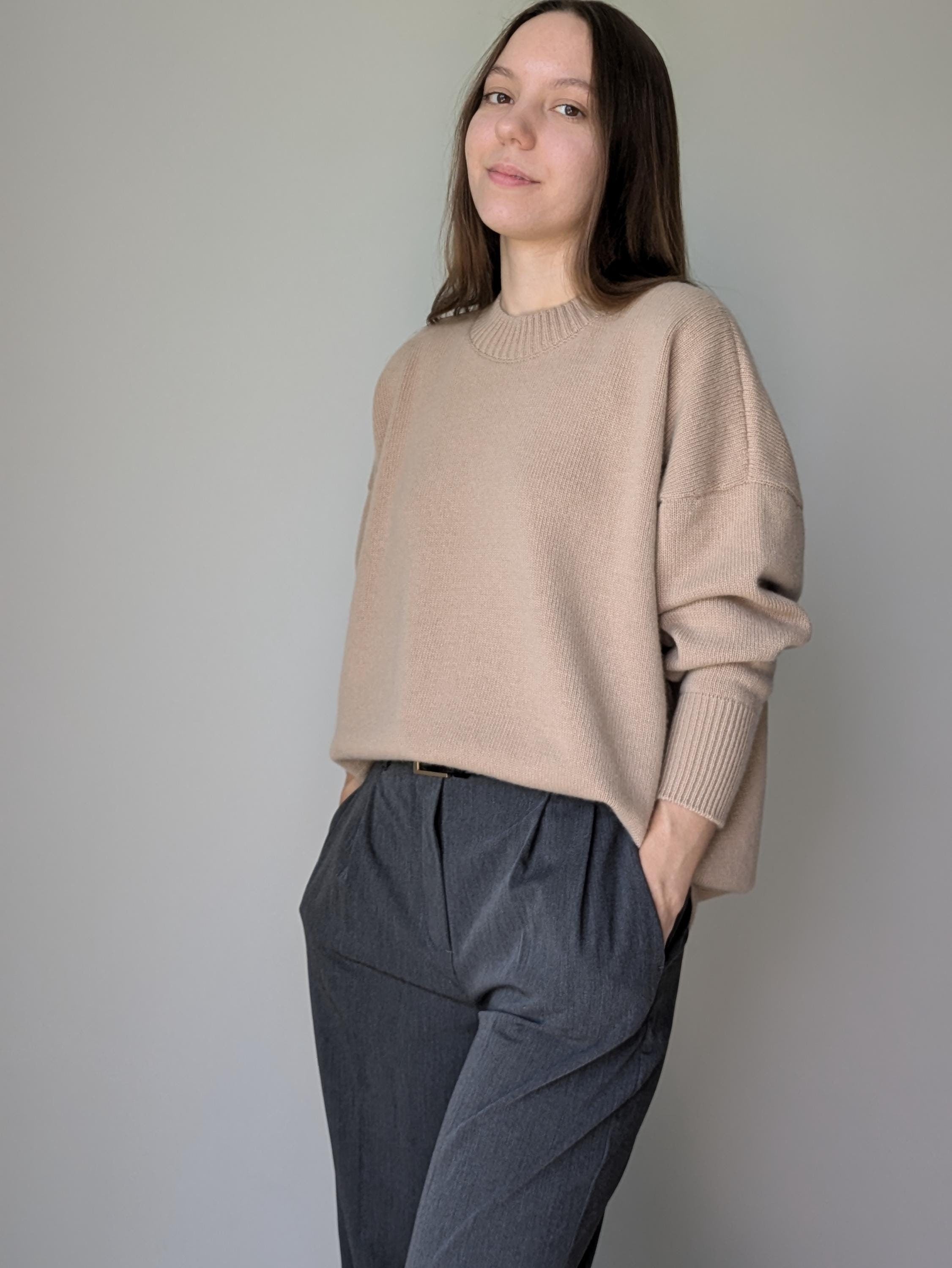 Woman wearing a beige sweater and dark gray pants against a plain background