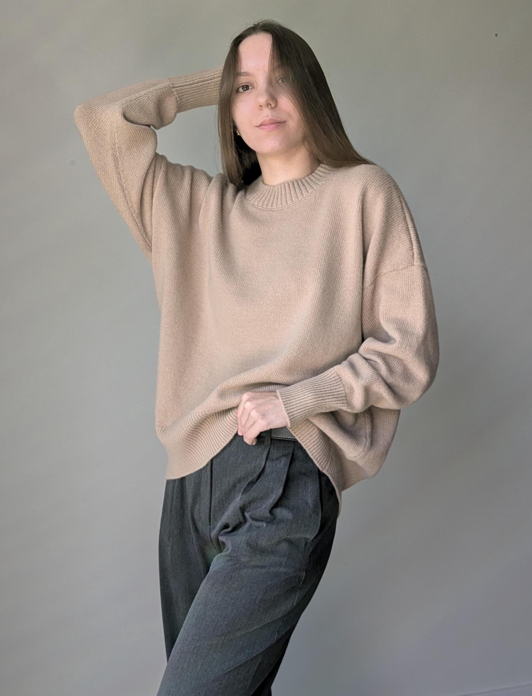 Person wearing a beige sweater and dark pants against a plain background