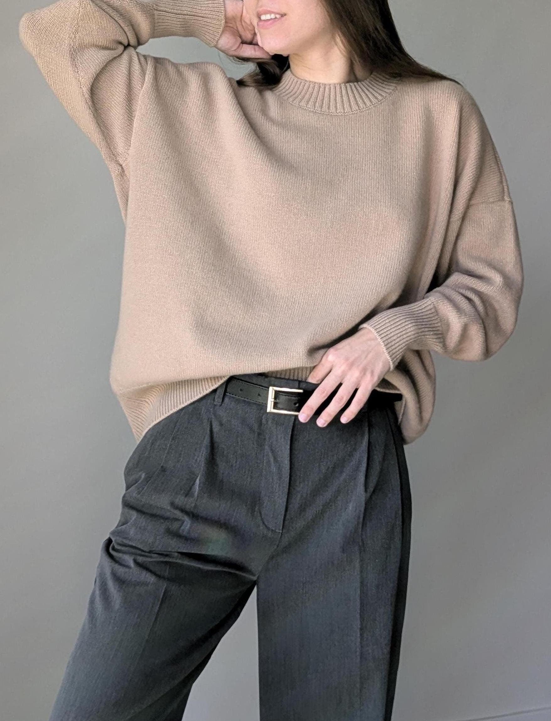 Person wearing a beige sweater and dark pants against a gray background