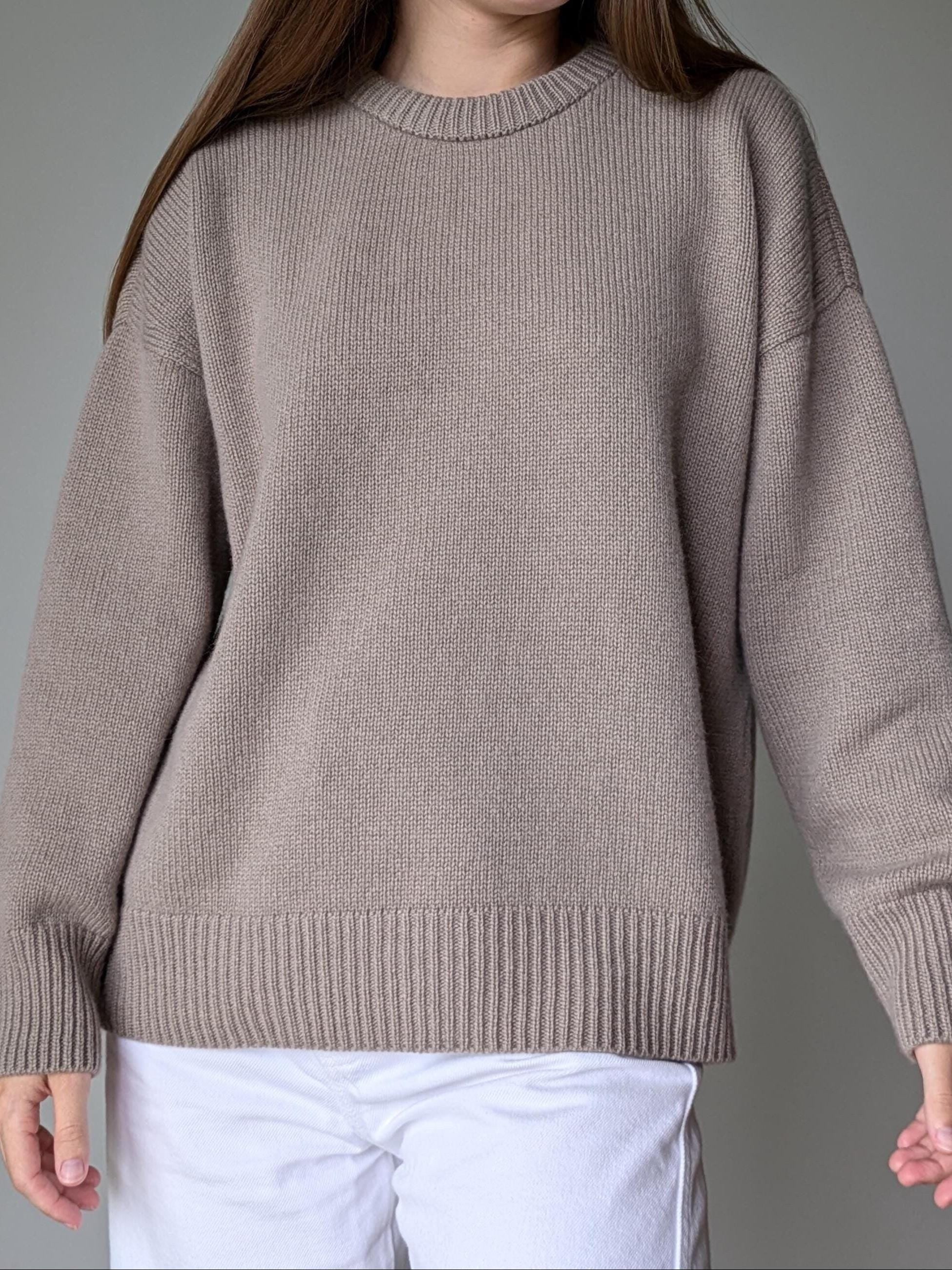 Person wearing a beige sweater and white pants against a gray background
