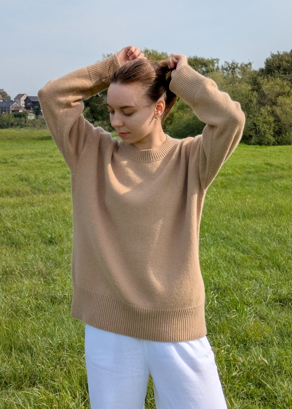Woman wearing a beige sweater and white pants standing in a grassy field.