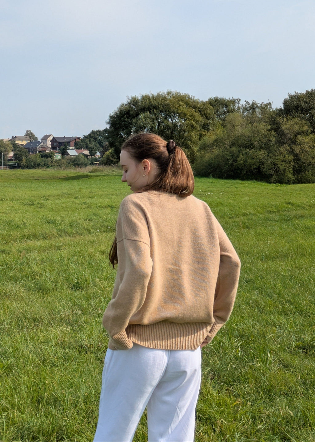 Person wearing a beige sweater and white pants standing in a grassy field with trees in the background