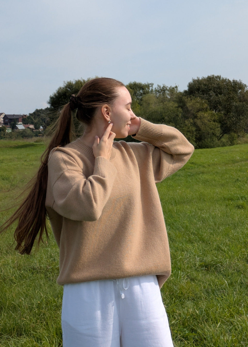 Woman in a beige sweater and white pants standing in a grassy field with trees in the background