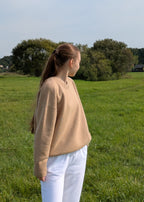 Person wearing a beige sweater and white pants standing in a grassy field with trees in the background