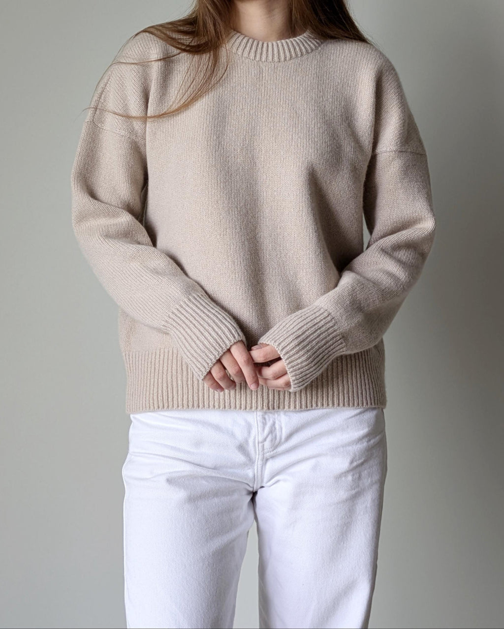 Person wearing a beige sweater and white pants against a gray background