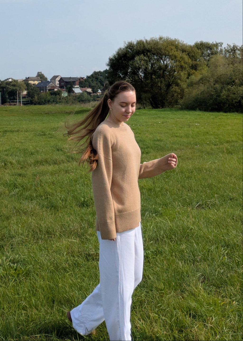 Woman walking in a field wearing a beige sweater and white pants.