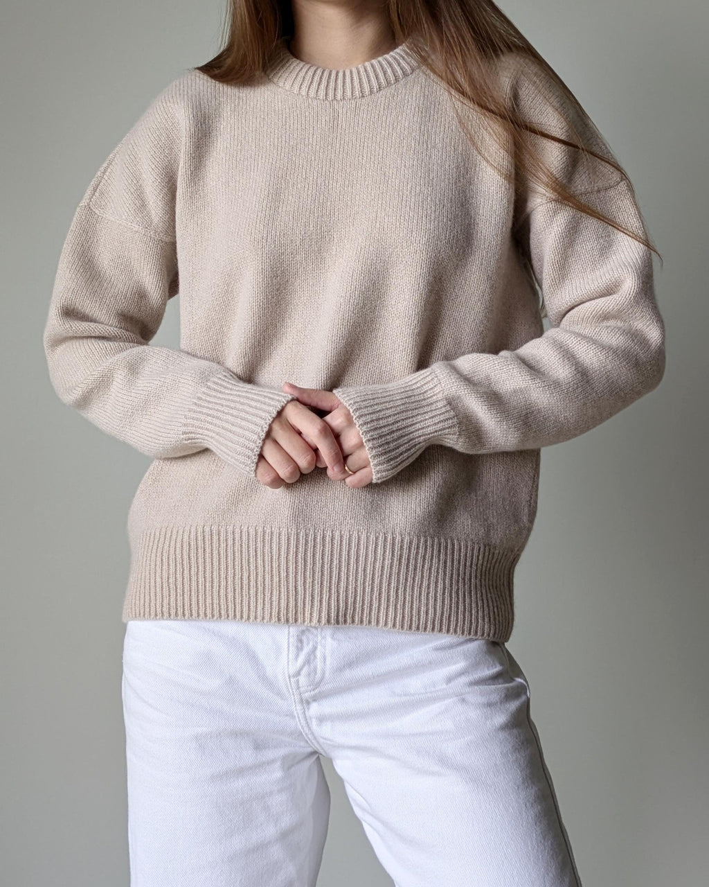 Person wearing a beige sweater and white pants against a gray background