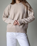 Person wearing a beige sweater and white pants against a gray background