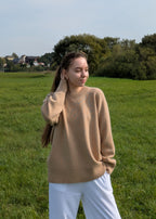 Woman wearing a beige sweater and white pants standing in a grassy field with trees in the background.