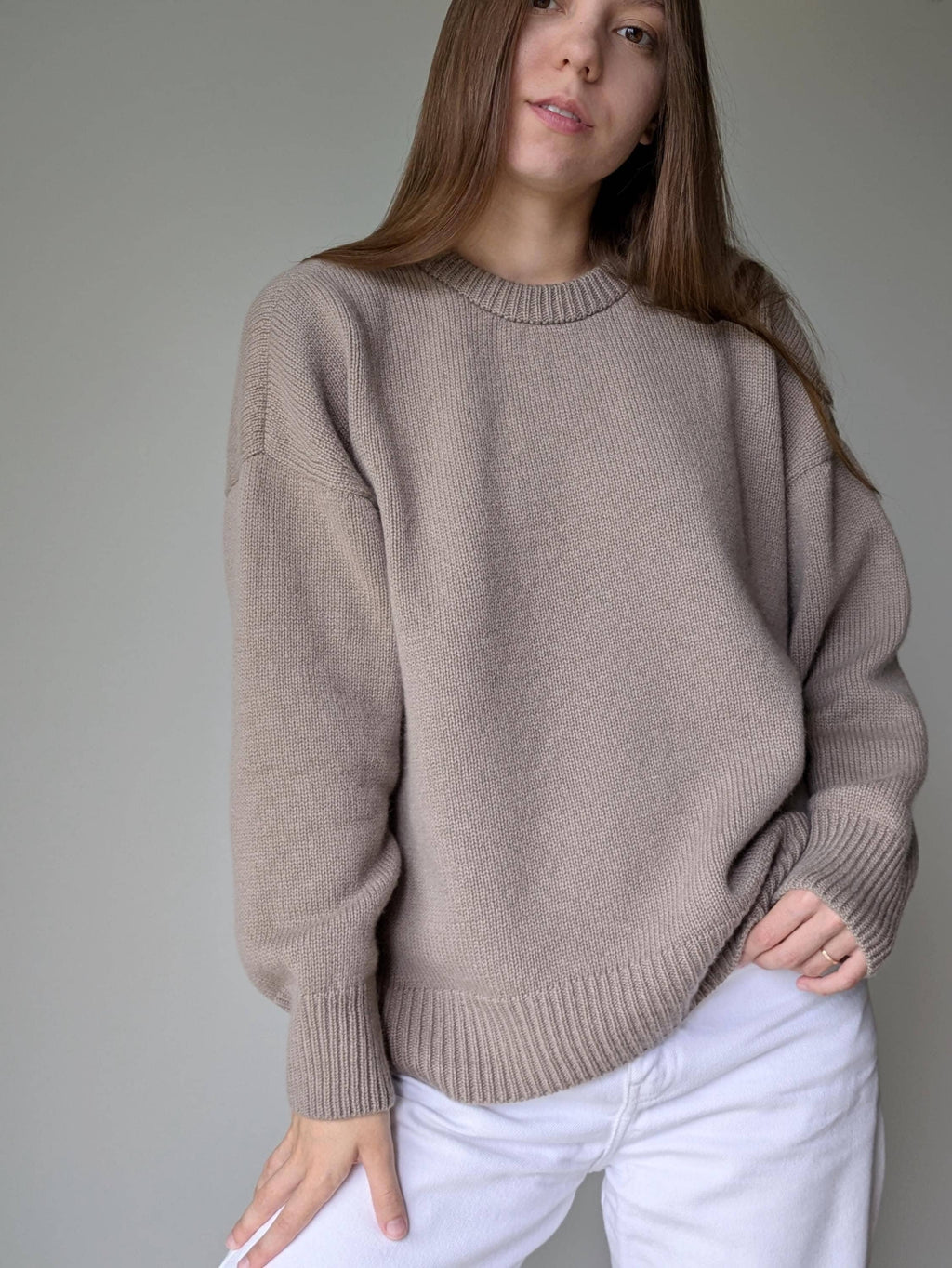 Woman wearing a beige sweater and white pants against a plain background