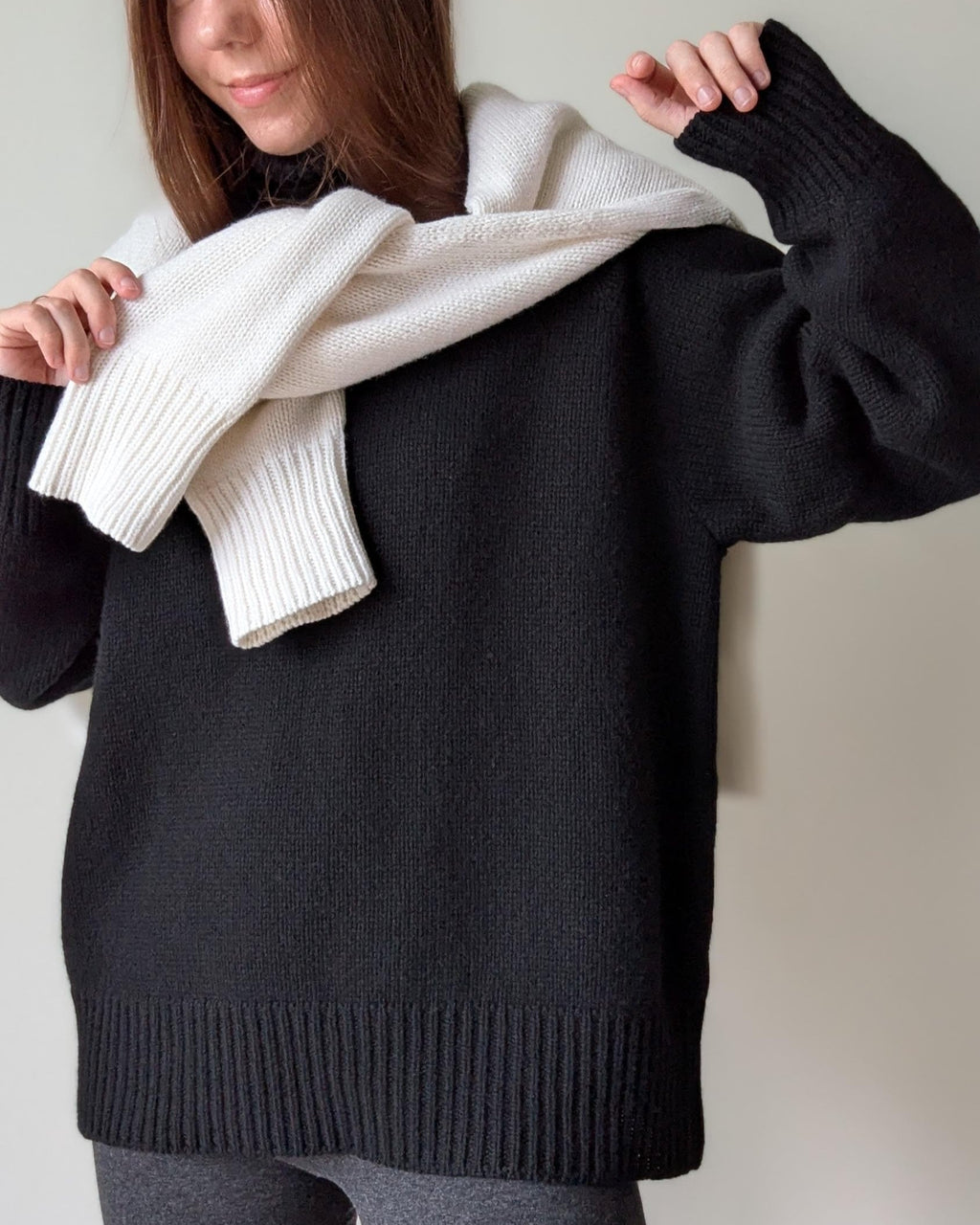 Black Person wearing a black sweater with a white scarf draped over their shoulders against a neutral background