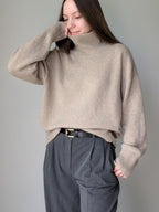 Beige Woman wearing a beige sweater and gray pants against a plain background
