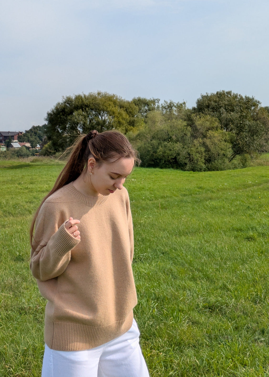 Woman in a beige sweater and white pants standing in a grassy field with trees in the background