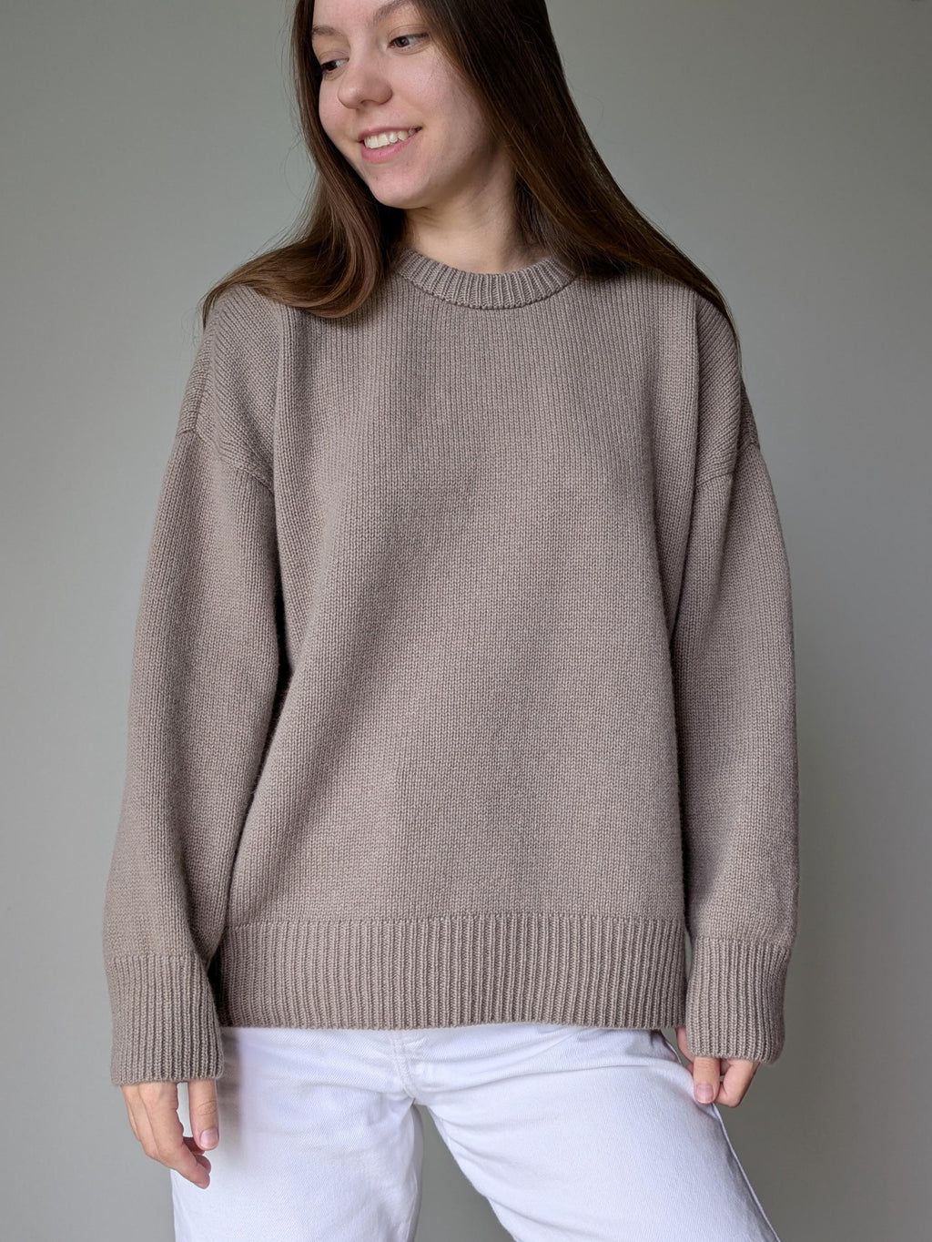 Woman wearing a beige sweater and white pants against a plain background