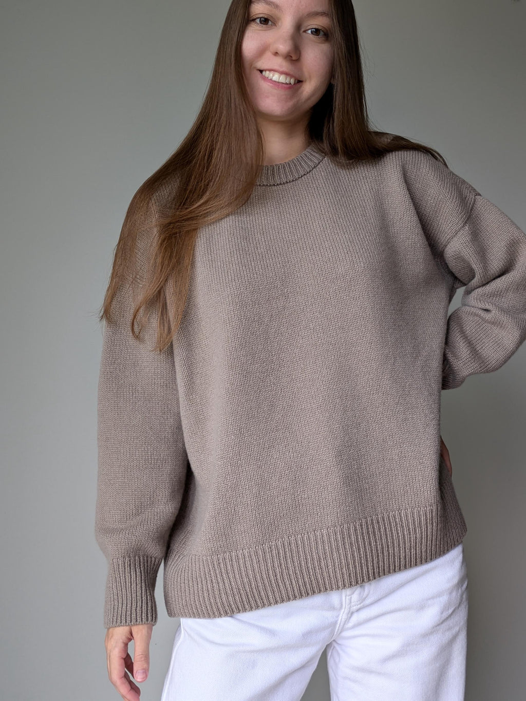 Woman wearing a beige sweater and white pants against a plain background